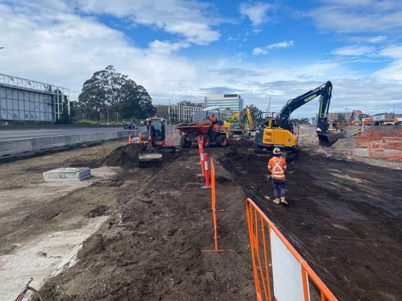 Sydney Gateway site photograph showing a civil infrastructure work area with road corridor, construction plant, traffic controls and workers in high visibility clothing.